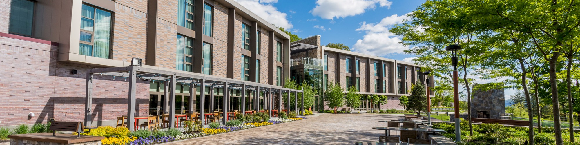 The image shows a modern building with large windows, outdoor seating, and a paved walkway. Trees and greenery line the area under a blue sky.