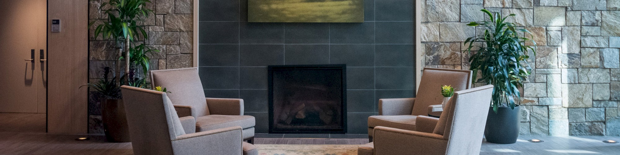 A cozy seating area with four armchairs, a rug, and a central fireplace with a landscape painting above it. The backdrop is a stone wall.