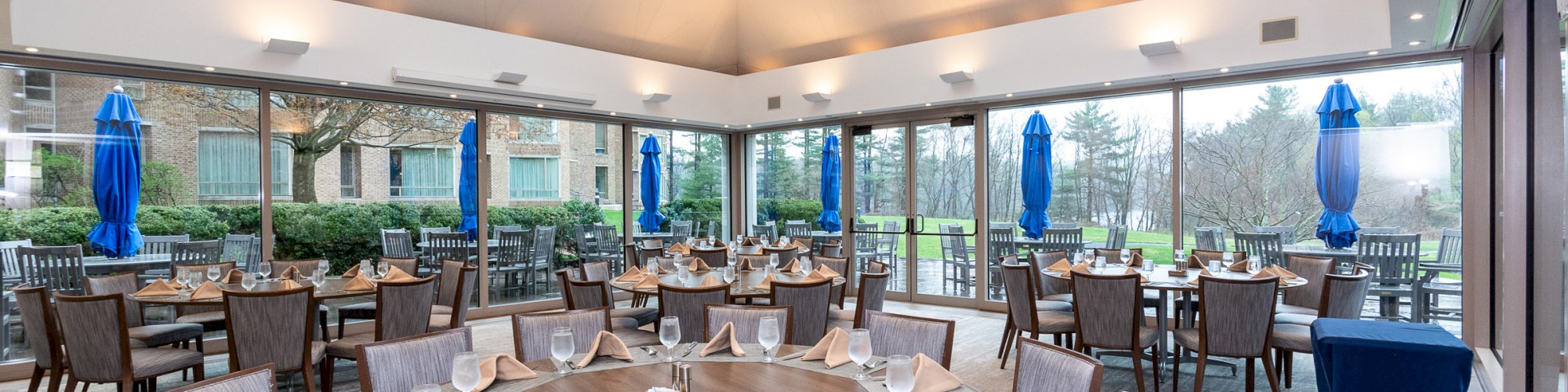 A restaurant dining area with a round table set for a meal, surrounded by chairs. Large windows reveal an outdoor patio with more seating.