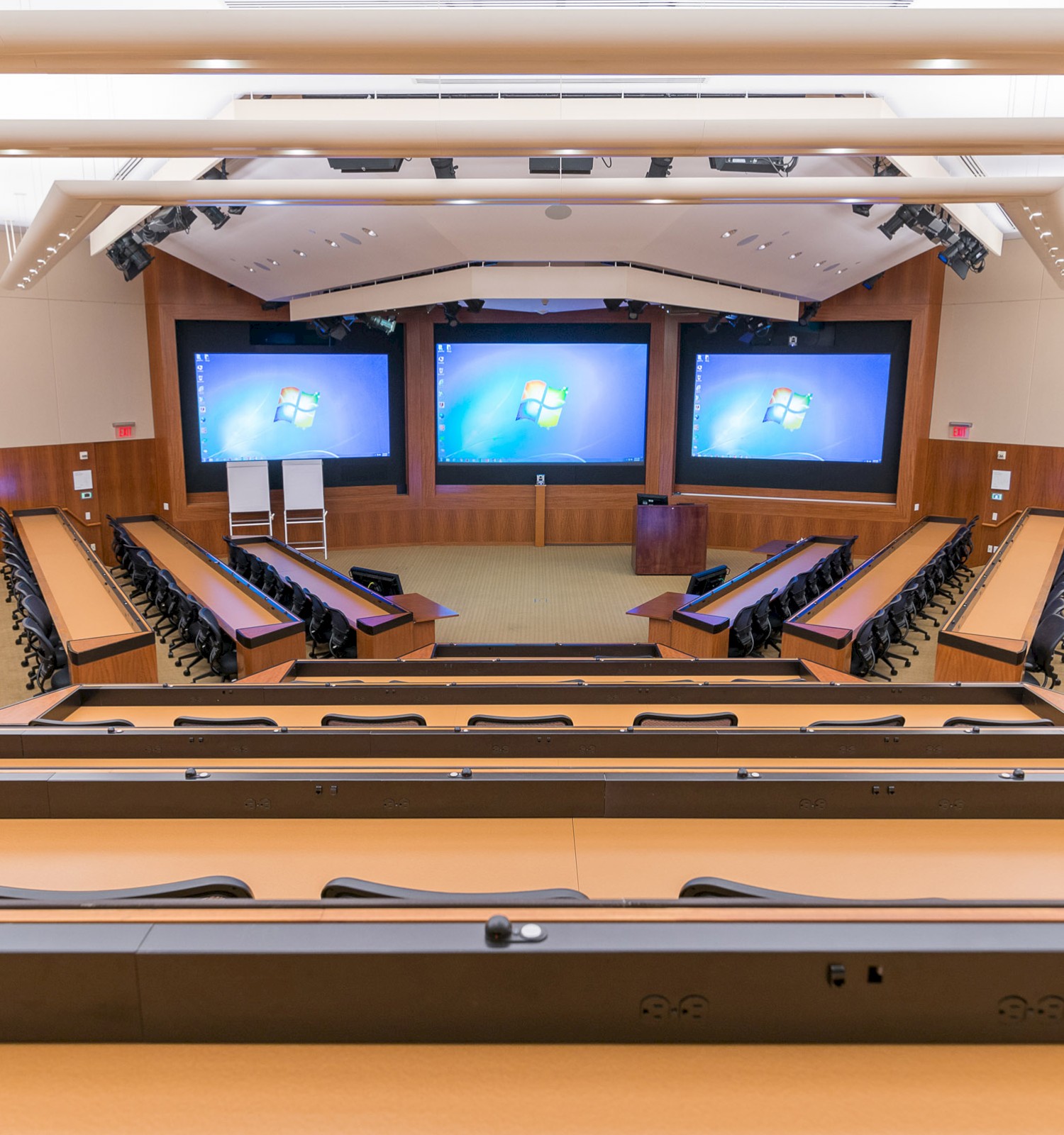 An empty, modern lecture hall with tiered seating, several computer screens at the front, and a central desk and whiteboards.