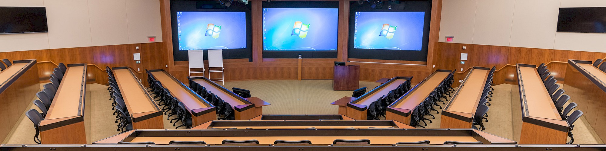 A modern lecture hall with tiered seating, multiple screens at the front displaying a Windows login screen, and a podium and whiteboards on the stage.