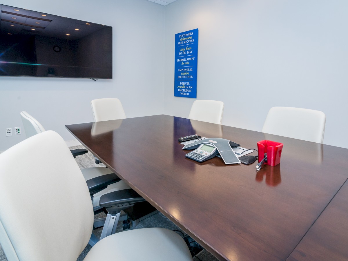 A conference room with a wooden table, white chairs, a wall-mounted television, and some items on the table, including a red cup.