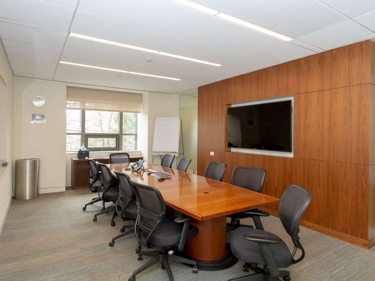 A modern conference room with a large wooden table, multiple office chairs, a whiteboard, a wall-mounted TV, and a window.