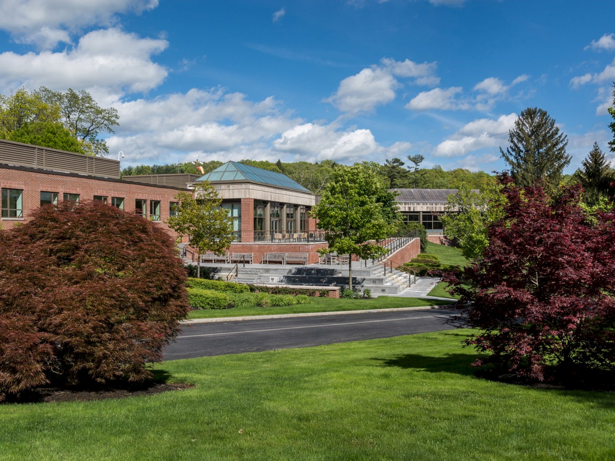 The image shows a picturesque, landscaped campus with green lawns, trees, and a modern building against a backdrop of a partly cloudy sky.