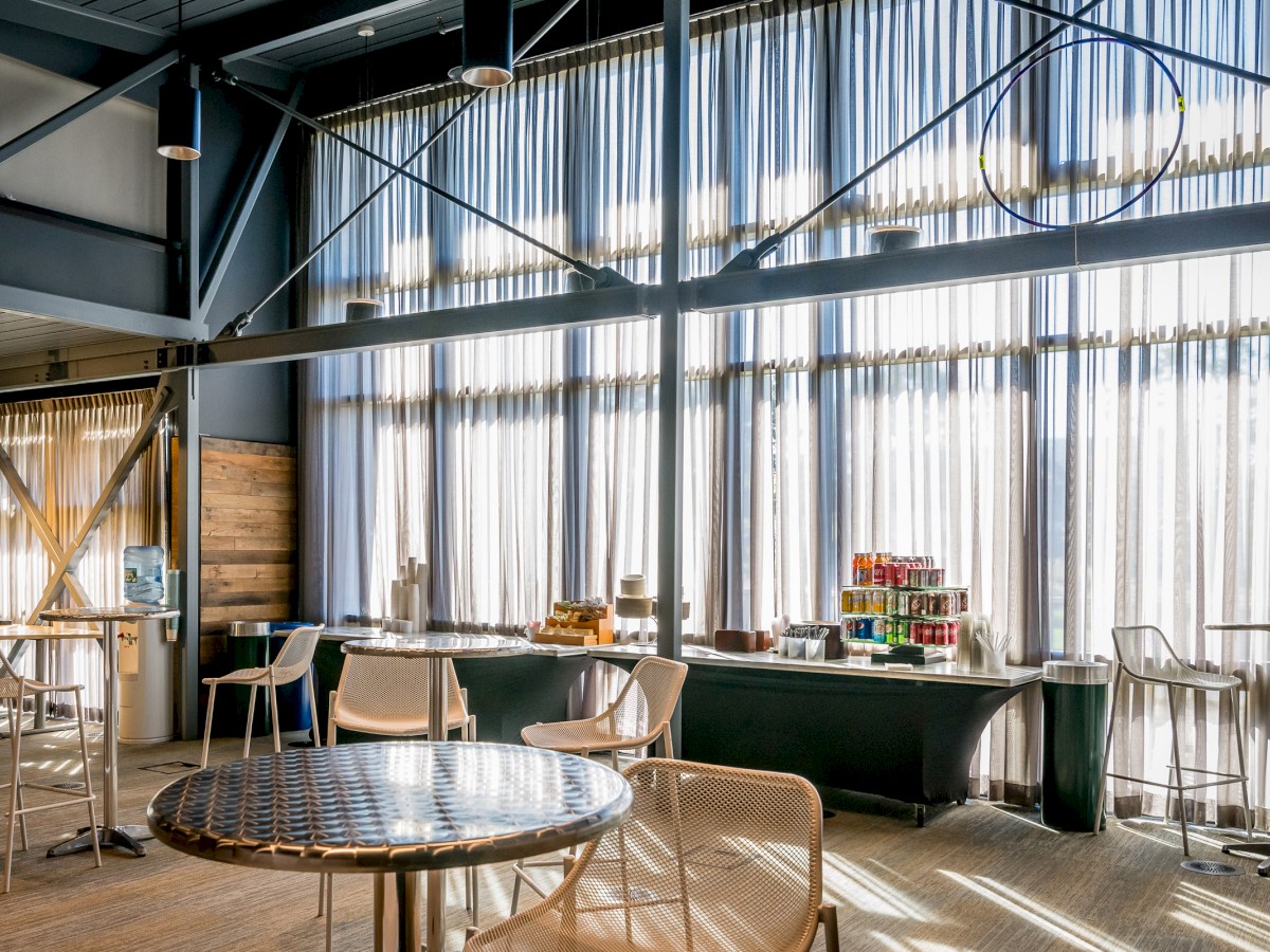 This image shows a stylish, sunlit cafeteria or break room with modern tables, chairs, and a refreshment area offering drinks and snacks.