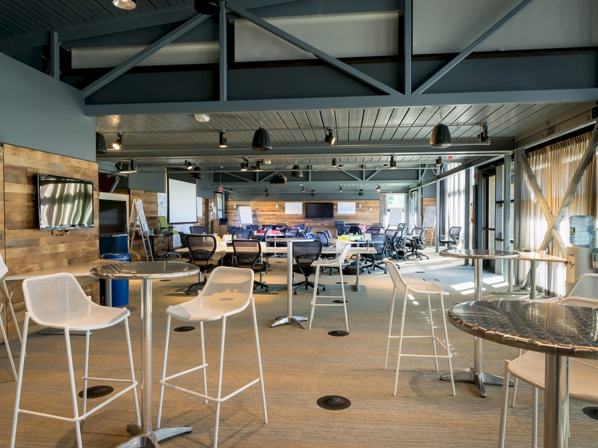 An open office space with high tables and stools, several desk stations, a TV, and industrial-styled decor.