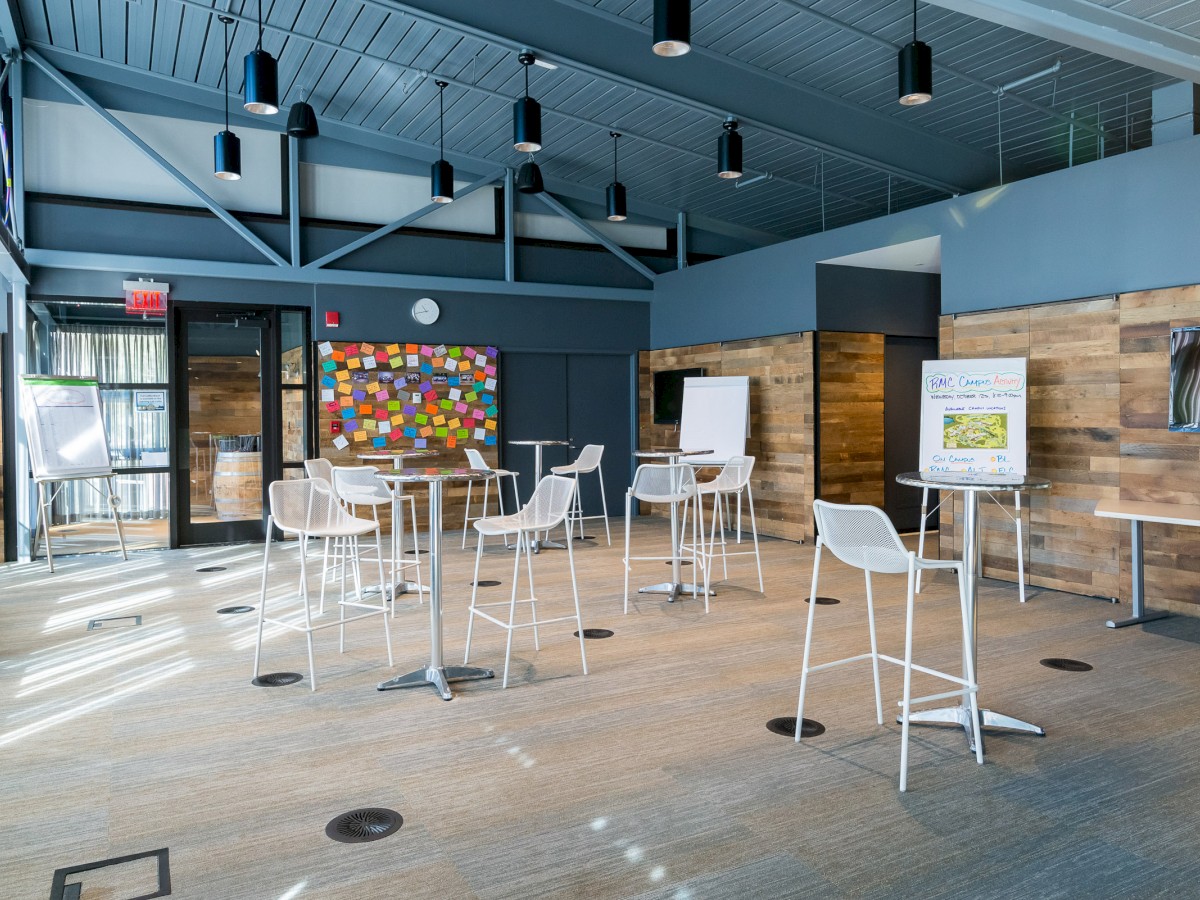 A modern meeting room with high ceilings, white chairs around small tables, flip charts, and colorful sticky notes on a bulletin board.