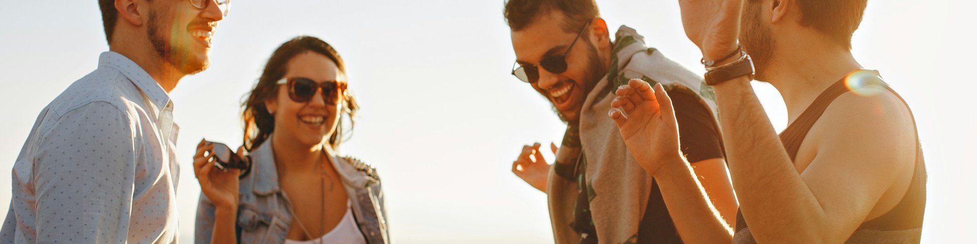 A group of four people, three men and one woman, are outdoors laughing and having a good time, with a clear sky in the background.