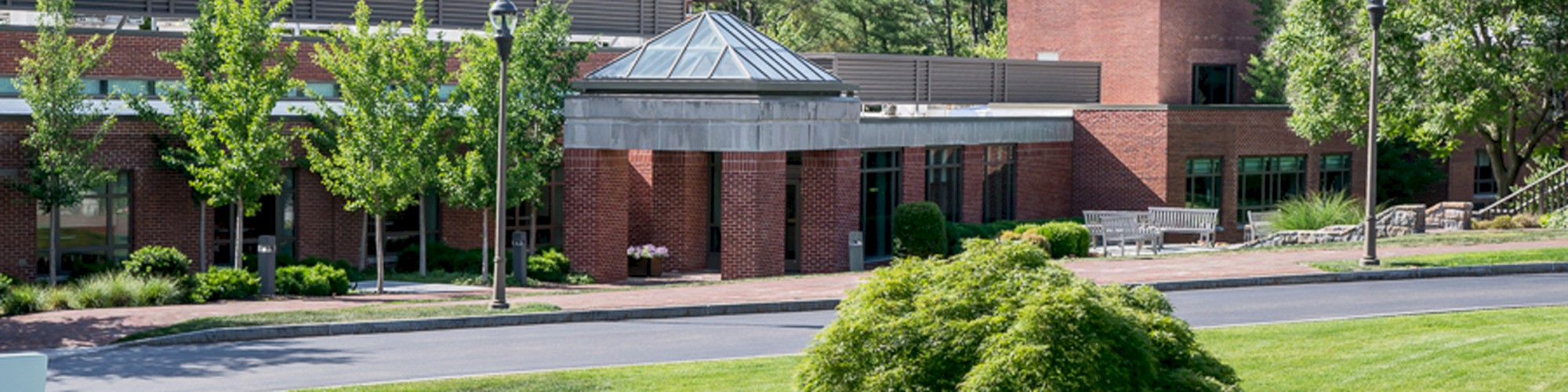 A brick building surrounded by green grass, trees, and shrubs, under a partly cloudy sky, with a driveway running in front and street lamps along the path.