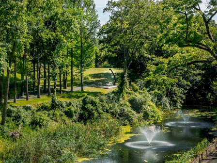 A scenic park with lush green trees, a winding path, and two water fountains in a pond under a partly cloudy sky.