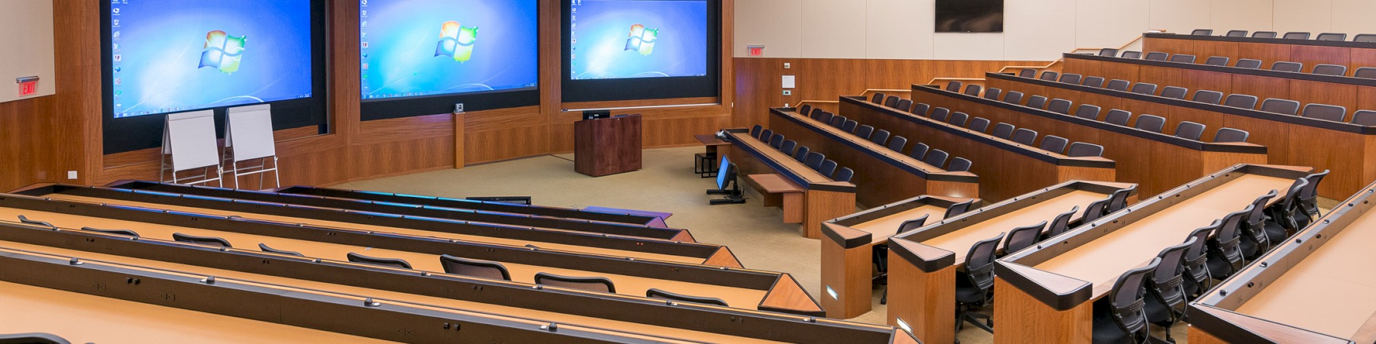 The image shows a large lecture hall with multiple screens at the front, several rows of seating, and a whiteboard.