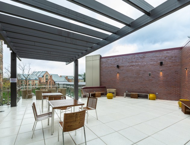 An outdoor patio area with tables and chairs under a pergola, next to a brick wall and stone building, with a scenic view in the background.