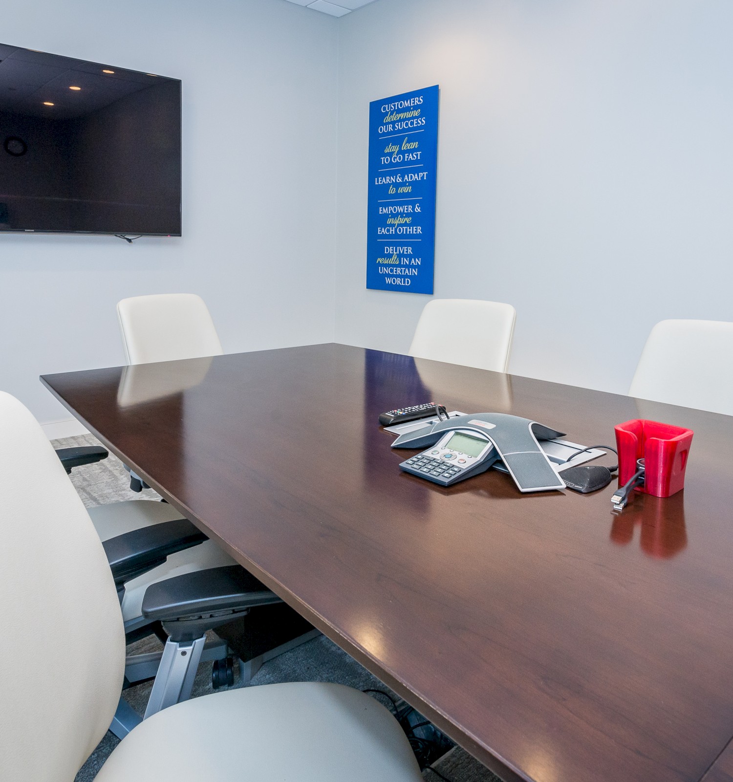 A conference room with a large table, white chairs, a wall-mounted TV, and items like a phone, cards, and a red cup on the table.