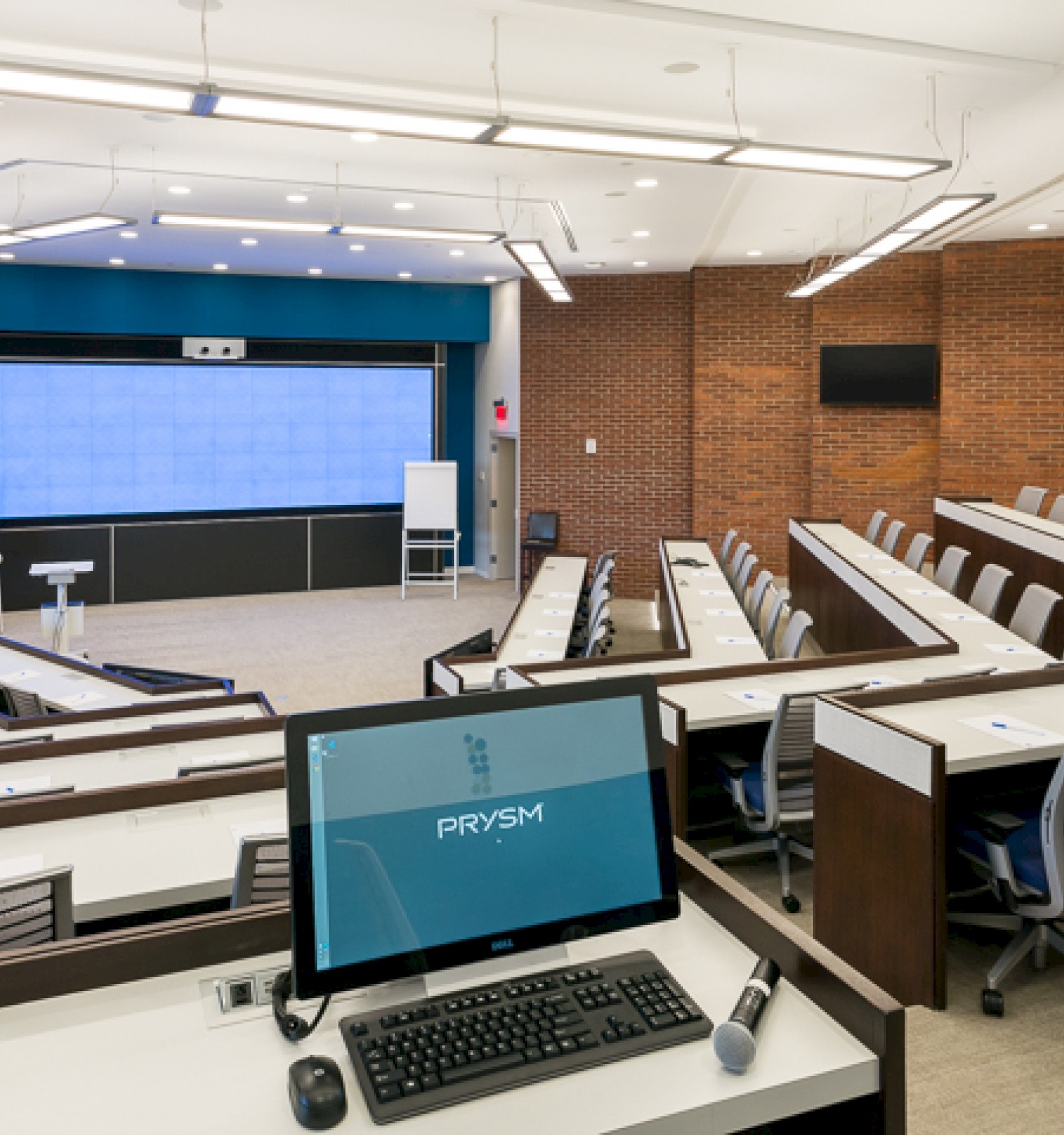 The image shows a modern lecture hall with tiered seating, a podium with a computer, and a large screen at the front.