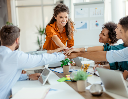 A group of people in a modern office setting, smiling and stacking hands in the center of a table, indicating teamwork and collaboration.