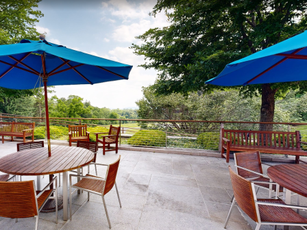 The image shows an outdoor patio area with wooden tables and chairs under blue umbrellas, surrounded by lush greenery.